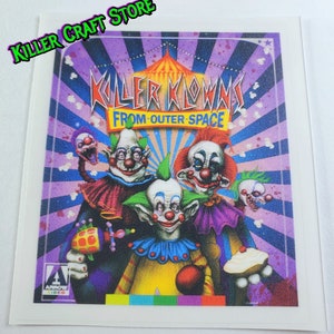 Könnte beinhalten: Ein Aufkleber mit einer bunten Illustration von vier Clowns mit dem Text "Killer Klowns from Outer Space". Der Aufkleber hat einen gestreiften Hintergrund in Lila, Blau und Weiß mit einem Stern oben rechts.