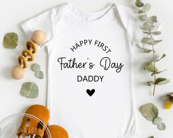 May include: White baby onesie with the text "Happy First Father's Day Daddy" and a small black heart. The onesie is surrounded by eucalyptus leaves, a wooden rattle, and a pacifier.