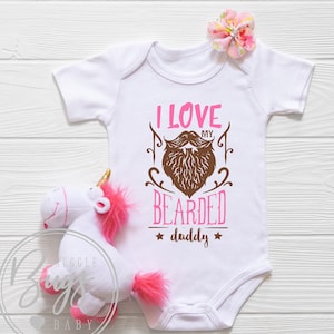 May include: White baby onesie with pink and brown text that reads "I love my bearded daddy". The onesie features a brown illustration of a beard with a pink mustache.