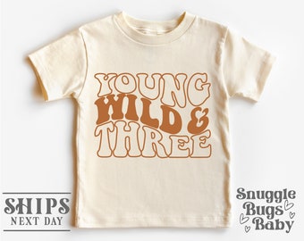May include: A beige short-sleeved t-shirt with the text "Young Wild & Three" in brown with a retro font style.