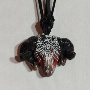 Könnte beinhalten: Eine schwarze Schnurkette mit einem schwarzen, roten und weißen Keramik-Anhänger in Form eines Widderkopfes mit einem einzigen Auge in der Mitte.