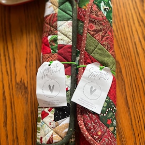 May include: Two rolled casserole hot pads with patchwork patterns in red, green, and white. Each pad has a tag that reads "Casserole Hot Pad" and "Handmade with Love." Care instructions are also printed on the tag.