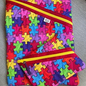 May include: A large quilted pouch and a smaller matching quilted pouch, both featuring a vibrant, multi-coloured puzzle piece pattern in red, yellow, blue, green, and purple. Each pouch has a red fabric border, a yellow zip, and a small white tag.