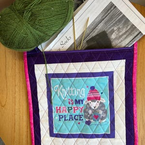 May include: A quilted knitting project bag with a purple zip, featuring a sheep graphic and the text "Knitting is my Happy Place." The bag is surrounded by a green ball of yarn, knitting needles, and a pattern.