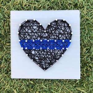 May include: A blue and black string art heart with a thin blue line across the center. The heart is made of string wrapped around nails hammered into a wooden board.