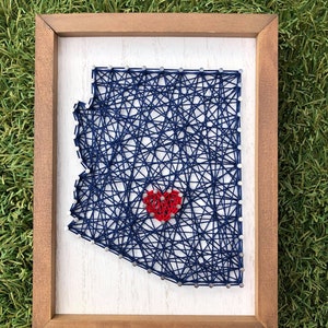 May include: A framed string art piece depicting the outline of Arizona with a red heart in the center. The string art is made with blue thread and silver nails.