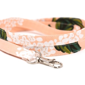 Könnte beinhalten: Ein pfirsichfarbenes Schlüsselband mit einem Blumenmuster aus weißen Blüten und grünen Blättern. Das Schlüsselband hat einen silberfarbenen Metallverschluss. Der Stoff ist mit weißen Blumen und grünen Blättern gemustert.