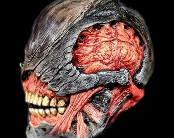 May include: A detailed, realistic, and gruesome skull mask with a cracked, gray, and black exterior revealing a red, fleshy, and bloody interior.