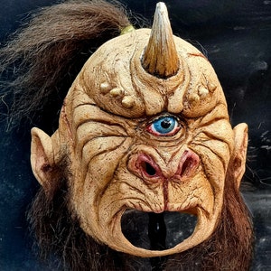 May include: A detailed, hand-painted mask of a creature with a single blue eye, a pointed horn, and a large, open mouth. The mask is made of a light brown material and has a textured surface.