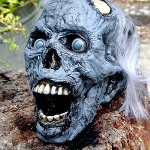 Peut inclure: Une tête de zombie grise, la bouche ouverte, avec des yeux bleus et des cheveux blancs. Le crâne a une apparence usée, avec des dents visibles et une texture en décomposition. Décoration d'Halloween.