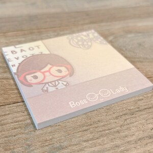 May include: A pink and beige notepad with a cartoon illustration of a woman wearing glasses and a white lab coat. The notepad has the text "Boss Lady" and a pair of glasses with eyelashes on the bottom.