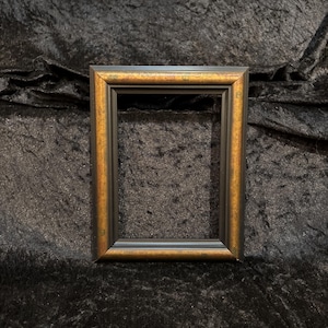 May include: A rectangular picture frame featuring a dark black outer edge and a warm, metallic gold inner border. The frame has a stepped, multi-layered profile with a smooth finish, designed to hold a photograph or artwork in a vertical orientation.