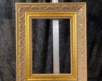 May include: An ornate gold picture frame with a rectangular aperture. The frame features a detailed, textured design with a lighter, neutral-toned border and a gold inner edge. The frame is displayed against a black backdrop.