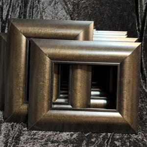 May include: A stack of rectangular picture frames featuring a dark bronze finish with a subtle metallic sheen. The frames have wide, beveled edges that slope inward toward the center opening, creating a layered, deep appearance in the stack.