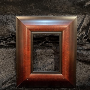 May include: A rectangular picture frame with a dark brown wood grain finish and a black inner border. The frame has a beveled edge and is set against a dark, textured background.