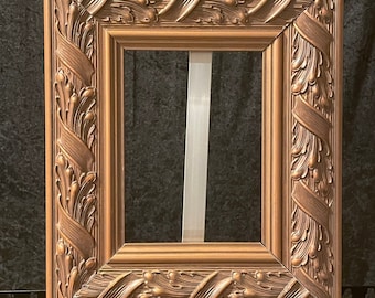 May include: A rectangular, ornate picture frame with a bronze finish. The frame features detailed relief carvings of stylized leaves and ribbons. The frame is displayed on a white easel against a dark background.