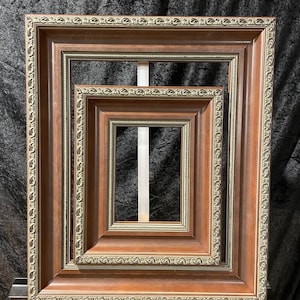 May include: Three nested, square picture frames with a dark brown finish and ornate gold-colored trim. The frames are displayed on a white easel against a black backdrop. The frames vary in size, creating a layered effect.