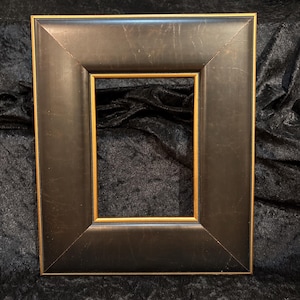 May include: Square picture frame with a dark brown finish and a gold inner edge. The frame has a beveled edge and is set against a dark, textured background. The frame is designed to hold a square image.