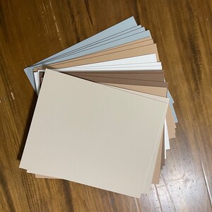 May include: A fan of various colored cardstock paper, including beige, brown, white, and gray. The papers are arranged on a wooden surface, showcasing a range of neutral tones. Ideal for crafting, scrapbooking, or other paper-based projects.