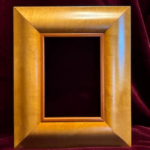 May include: A rectangular picture frame with a gold finish. The frame has a wide, beveled edge and a narrower inner frame. The background is a dark burgundy color.