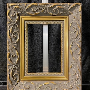 May include: A square picture frame with ornate, gold-colored floral and scrollwork details. The frame has a gray outer border and a gold inner border. The frame is empty, with no picture inside.