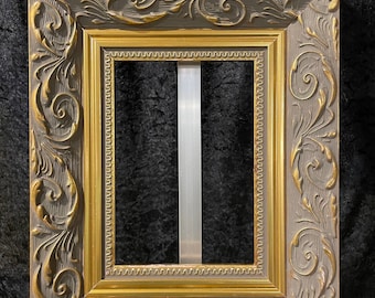 May include: A square picture frame with ornate, gold-coloured floral and scrollwork details. The frame has a grey outer border and a gold inner border. The frame is empty, with no picture inside.
