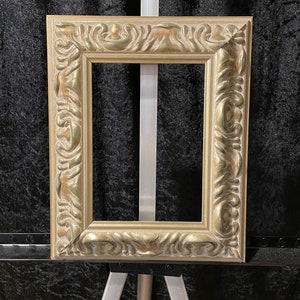 May include: Ornate, rectangular picture frame with a gold finish and detailed scrollwork. The frame is empty, showcasing its decorative design. The frame is displayed on a silver easel against a black backdrop.