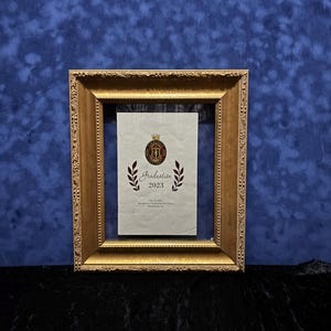 May include: A gold-colored framed document with the text "Graduation 2023" and a crest. The frame has ornate detailing and a dark background. The document is centered within the frame.