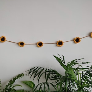 May include: A garland of crocheted sunflowers strung on twine, hanging on a white wall. The garland is decorated with six yellow sunflowers with brown centers.