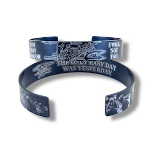 May include: Two dark blue metal cuff bracelets. One bracelet is on top of the other. The top bracelet has the text "I AM THAT WARRIOR" and "I WILL NOT FAIL". The bottom bracelet has the text "THE ONLY EASY DAY WAS YESTERDAY".