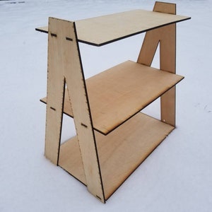 May include: A three-tiered wooden shelf unit with a triangular frame. The shelf is made of light-colored wood and has a natural finish.
