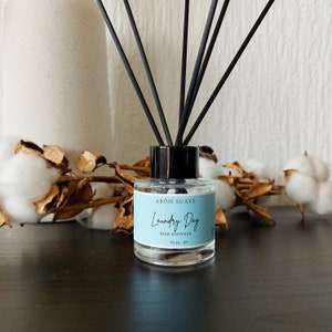Op de afbeelding: Een heldere glazen fles met een zwarte dop en zwarte rietjes. De fles bevat een rietjesdiffuser met het label "Arom Suave Laundry Day Reed Diffuser 50 ml e".
