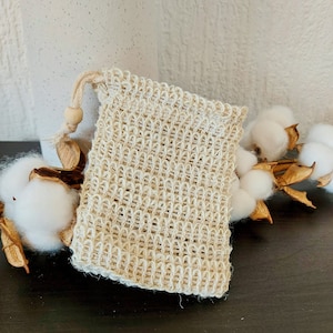 May include: A close-up of a small, beige, crocheted drawstring bag made of natural fibers. The bag is sitting on a dark surface with a sprig of cotton flowers in the background.