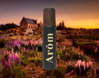 May include: A black bottle with a wood-grain cap and the word "Arom" in gold lettering. The bottle is in front of a field of purple flowers and a stone building in the background.