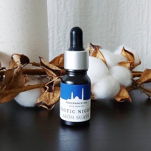 Op de afbeelding: Een klein zwart flesje met een zilveren dop en een blauw etiket met de tekst "FRAGRANCE OIL HIGH QUALITY EXOTIC NIGHT AROM SUAVE".