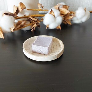 Peut inclure: Un pain de savon carré de couleur lavande repose sur un petit plateau rond en bambou tressé. Le savon est texturé et semble fait à la main. En arrière-plan, une branche de boules de coton et de gousses séchées ajoute un élément naturel.