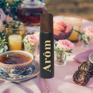 Könnte beinhalten: Eine schwarze Flasche mit einem Holzverschluss und dem Wort "Arom" in goldenen Buchstaben. Die Flasche steht auf einer rosa Tischdecke mit einer Tasse Tee und Blumen im Hintergrund.