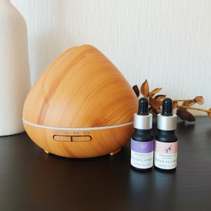 Op de afbeelding: Een houten look etherische olie diffuser met twee kleine flesjes geurolie. De flesjes zijn gelabeld met "Wildflower" en "Fresh Flower". De diffuser heeft licht- en mistinstellingen.