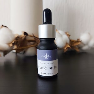 Op de afbeelding: Een klein zwart flesje met een zilveren dop en een druppelaar. Het flesje heeft een etiket met de tekst "Fragrance Oil Cedar & Amber Arom Suave".
