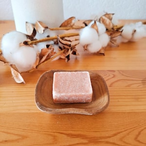 May include: A square, light pink soap bar rests on a small, wooden, oval-shaped dish. The dish sits on a wooden surface, with cotton stems and a white vase in the background. The soap bar is the focal point of the image.
