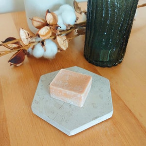 Puede incluir: Una pastilla de jabón cuadrada de color naranja claro descansa sobre un jabonera hexagonal gris claro. La jabonera tiene un aspecto marmóreo. El fondo incluye una botella de vidrio verde, tallos de algodón y una superficie de madera.