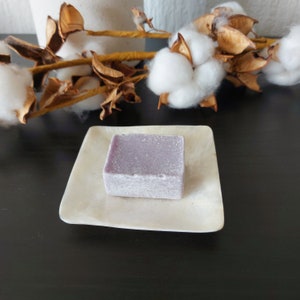 Op de afbeelding: Een kleine, vierkante, lavendelkleurige zeepstaaf ligt op een wit, ovaalvormig schaaltje. Het schaaltje is gemaakt van een natuurlijk materiaal, mogelijk schelp of bot.