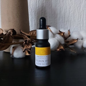 Op de afbeelding: Een klein zwart flesje met een zilveren dop en een geel etiket. Het etiket vermeldt "FRAGRANCE OIL HIGH QUALITY GOLDEN HOUR AROM SUAVE".