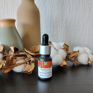 Puede incluir: Un pequeño frasco negro con tapa plateada y gotero. El frasco tiene una etiqueta colorida que dice "Fragrance Oil High Quality Patchouli Arom Suave".