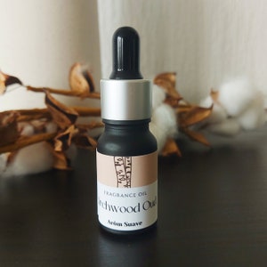 Op de afbeelding: Een klein zwart flesje met een zilveren dop en een druppelaar. Het flesje heeft een etiket met de tekst "Fragrance Oil" en "Teakwood Oud" in zwarte tekst op een perzikkleurige achtergrond. Het etiket heeft ook de tekst "Arom Suave" in zwarte tekst op een witte achtergrond.