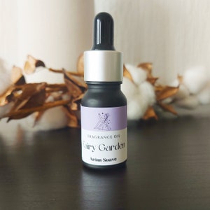 Puede incluir: Un pequeño frasco de vidrio negro con tapa plateada y gotero. El frasco tiene una etiqueta que dice "Fragrance Oil Fairy Garden Arom Suave".