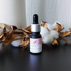 Op de afbeelding: Een klein zwart flesje met een zilveren dop en een druppelaar. Het flesje heeft een roze etiket met de tekst "FRAGRANCE OIL HIGH QUALITY FRESH FLOWERS AROM SUAVE".