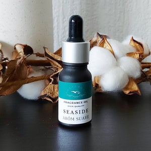 Op de afbeelding: Een klein zwart flesje met een zilveren dop en een groen label waarop "Fragrance Oil High Quality Seaside Arom Suave" staat. Het flesje staat op een donker oppervlak met gedroogde katoenbollen op de achtergrond.