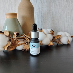 May include: A small black glass bottle with a silver cap and a dropper. The bottle is labeled "Fragrance Oil High Quality Leche de Coco Arom Suave". The bottle is sitting on a dark wooden surface with cotton bolls and dried branches.