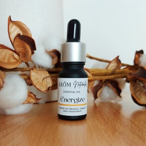Op de afbeelding: Een zwarte glazen fles met een zilveren dop en een zwarte druppelaar. De fles is gelabeld met "ARÔM Naturals Essentiële Olie Energize Mengsel van Oranje, Citroen en Grapefruit".